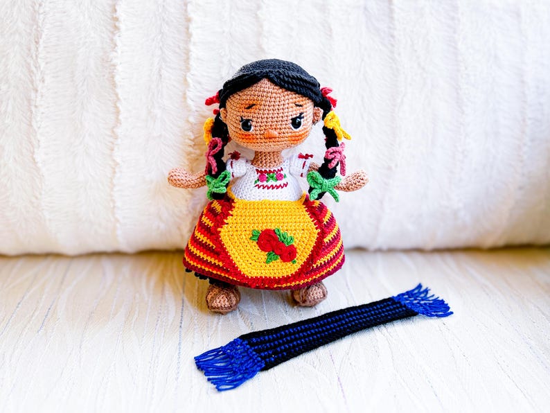 A crochet doll wears Purépecha clothing, including an embroidered white blouse and gold apron, a striped skirt and brown sandals. Her black hair is in two braids adorned with colorful ribbons. A blue and black rebozo lays spread out in front of her.