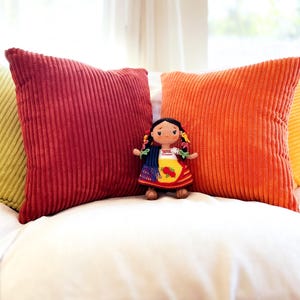 A crochet Mexican doll dressed in Purépecha attire stands on a sofa surrounded by pillows. She wears a white blouse, striped skirt and gold apron. Her hair is braided and adorned with colorful ribbons. A blue and black rebozo hangs over her shoulder.