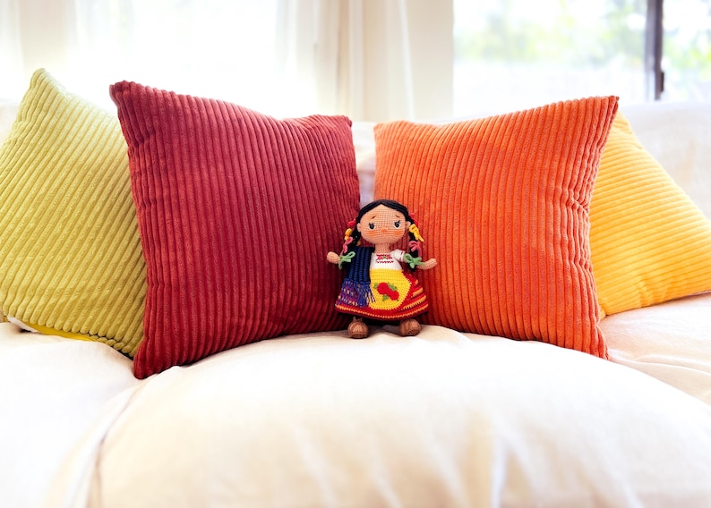 A crochet Mexican doll dressed in Purépecha attire stands on a sofa surrounded by pillows. She wears a white blouse, striped skirt and gold apron. Her hair is braided and adorned with colorful ribbons. A blue and black rebozo hangs over her shoulder.