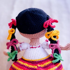 A crochet Mexican doll is seen from behind. Her black hair is in two braids adorned with colorful ribbons. She wears a white blouse with embroidery and a striped skirt.