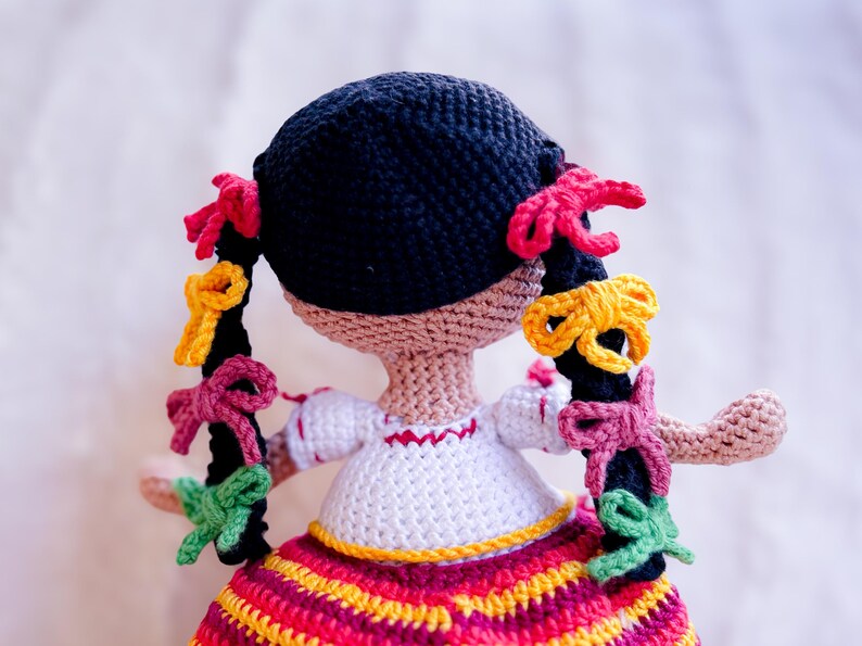 A crochet Mexican doll is seen from behind. Her black hair is in two braids adorned with colorful ribbons. She wears a white blouse with embroidery and a striped skirt.