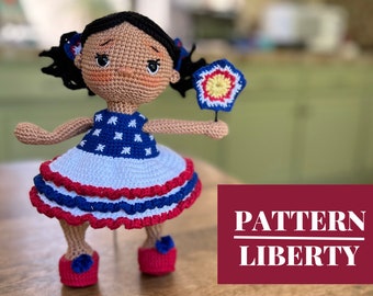 Può includere: Bambola all'uncinetto con pelle marrone, capelli neri e un vestito rosso, bianco e blu. La bambola tiene un oggetto a forma di stella rosso, bianco e blu. Il testo "PATTERN LIBERTY" è visibile nell'immagine.