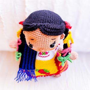 A crochet amigurumi doll is seen from above dressed in a style indigenous to Michoacán, Mexico. Her black hair is in two braids with colorful ribbons. She wears an embroidered blouse and gold apron. A blue and black rebozo hangs over her shoulder.