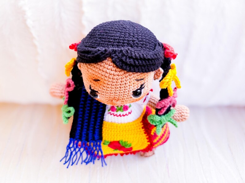 A crochet amigurumi doll is seen from above dressed in a style indigenous to Michoacán, Mexico. Her black hair is in two braids with colorful ribbons. She wears an embroidered blouse and gold apron. A blue and black rebozo hangs over her shoulder.