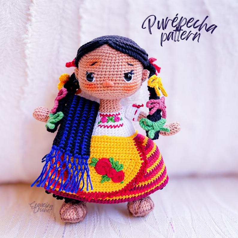 A crochet Mexican doll is dressed in clothing indigenous to the Purépecha of Michoacán. Her black hair is in braids with colorful ribbons. She wears an embroidered blouse, gold apron and striped skirt. A blue and black rebozo hangs over her shoulder.