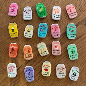 May include: A collection of miniature, hand-painted Olipop soda can replicas. Each can features a different flavor name, such as "Cherry Vanilla" and "Orange Cream", and colorful designs. The cans are arranged on a wooden surface.
