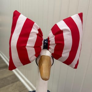 May include: A large fabric bow featuring vertical red and white stripes with a dark blue center knot patterned with small white stars. The bow is attached to a headband worn by a white goose figurine that is wearing a dark blue star-patterned outfit.