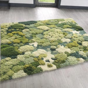 Puede incluir: Una alfombra rectangular con un diseño texturizado similar al musgo. La alfombra presenta una variedad de tonos verdes, incluyendo verde claro, verde oscuro y blanco roto, creando una apariencia natural y orgánica. El diseño de la alfombra se asemeja a un paisaje de pequeñas colinas redondeadas.