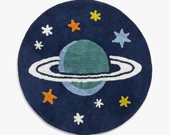 May include: A circular navy blue rug featuring a central teal planet with a white ring. The design includes scattered stars in yellow, orange, and white, along with a light blue star. The rug has a textured, tufted surface throughout the design.