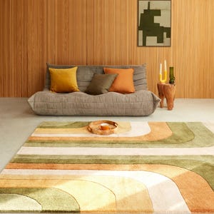 May include: A living room scene featuring a gray sofa with colorful throw pillows in yellow, olive, and orange. A patterned rug in shades of green, orange, and cream dominates the foreground. A wooden tray with a cup sits on the rug, and a wooden side table holds candles.