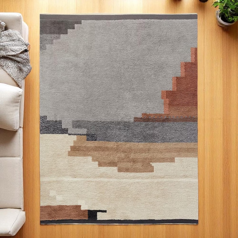May include: A rectangular rug with a geometric pattern in grey, brown, and cream. The design includes a stepped pattern in the upper right corner and a border along the top edge. The rug is placed on a wooden floor.