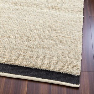 May include: Cream-coloured, textured rug with a dark grey border and a cream-coloured trim. The rug has a thick, plush pile, and is displayed on a dark wood floor. The rug's texture is visible.