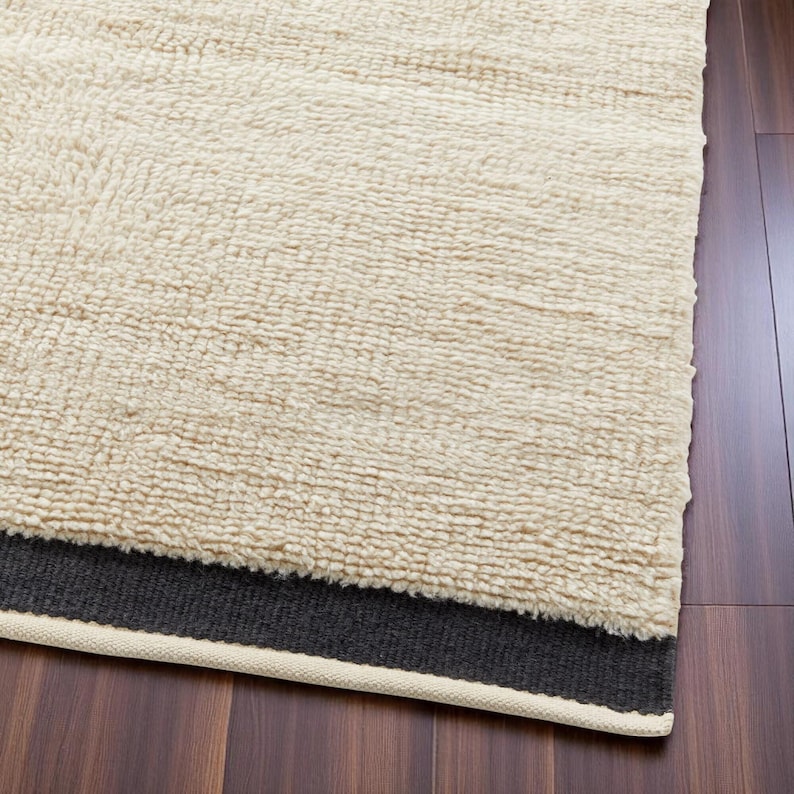 May include: Cream-coloured, textured rug with a dark grey border and a cream-coloured trim. The rug has a thick, plush pile, and is displayed on a dark wood floor. The rug's texture is visible.