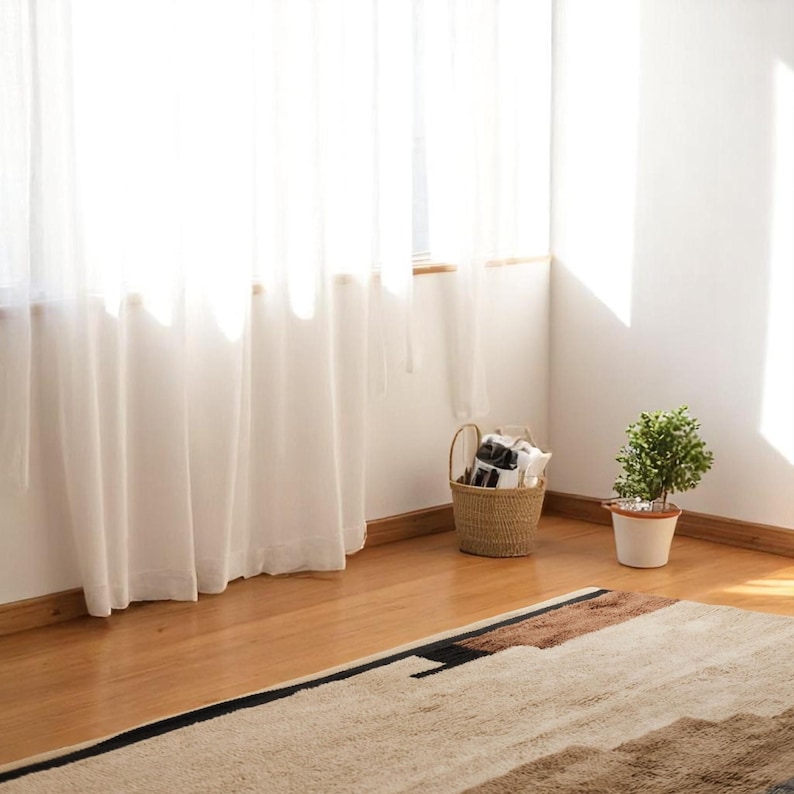 May include: A bright interior corner featuring white sheer curtains, a wicker basket, and a potted plant. A beige rug with black and brown geometric patterns sits on the wooden floor, creating a cosy atmosphere.