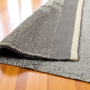 May include: A rolled-up grey rug with a textured surface, revealing a section of the underside. The rug has a border of a lighter colour and is placed on a wooden floor.