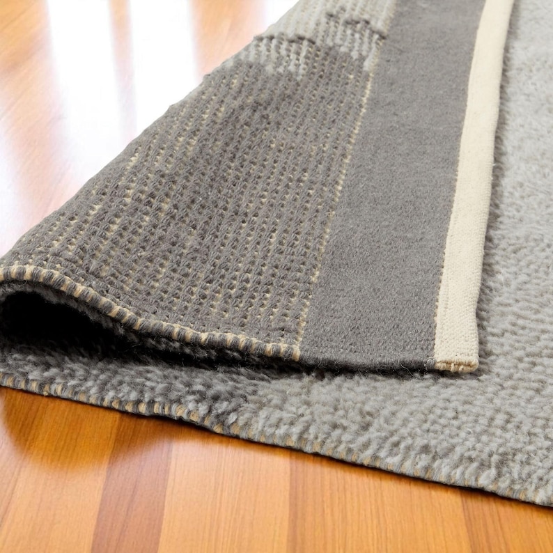 May include: A rolled-up grey rug with a textured surface, revealing a section of the underside. The rug has a border of a lighter colour and is placed on a wooden floor.