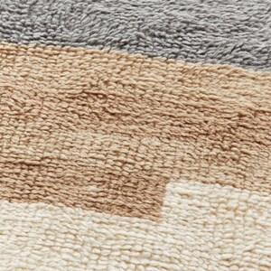 May include: Close-up of a textured rug with horizontal stripes in shades of grey, tan, brown, and cream. The rug has a plush, soft appearance, with a dense, looped pile. The colour palette is neutral and earthy.