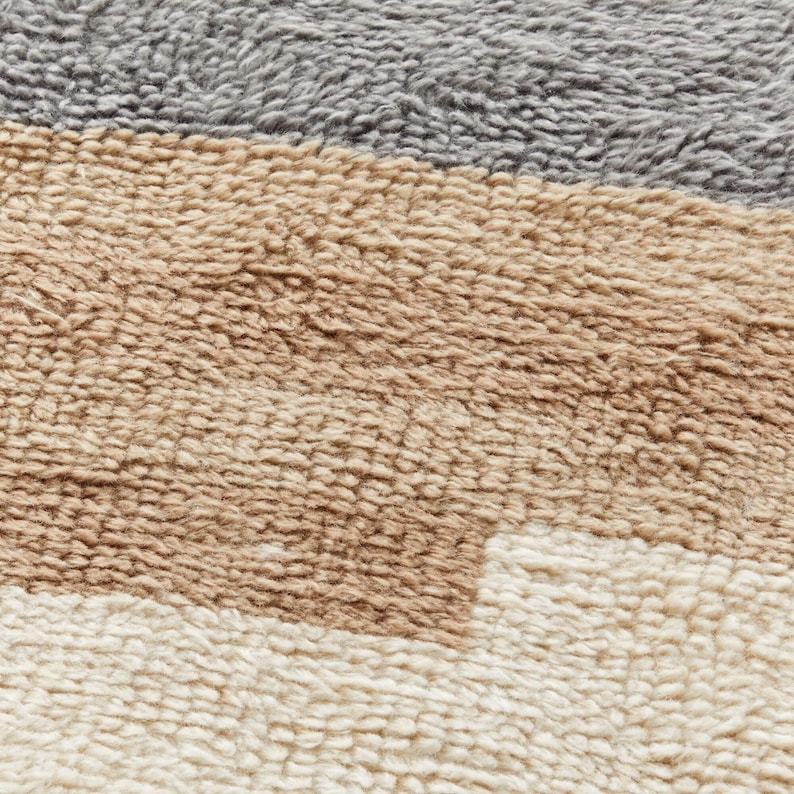 May include: Close-up of a textured rug with horizontal stripes in shades of grey, tan, brown, and cream. The rug has a plush, soft appearance, with a dense, looped pile. The colour palette is neutral and earthy.