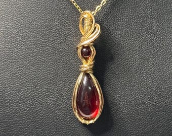 May include: A gold-colored pendant necklace featuring a teardrop-shaped, deep red gemstone. The gemstone is encased in a gold wire frame, with a smaller red bead above it. The necklace chain is also gold.