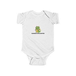 May include: White baby bodysuit with a cartoon dragon graphic. The green dragon has orange horns and a red tail. The text "RANDOM ENCOUNTER" is printed below the dragon in black. The bodysuit is made of soft material.
