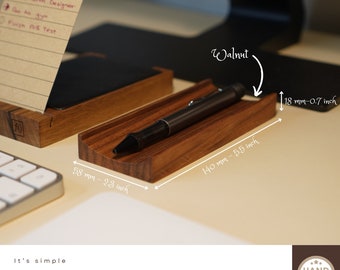 May include: A wooden pen holder made of walnut wood. It measures 140 mm long, 58 mm wide, and 18 mm tall. The pen holder is sitting on a white surface.