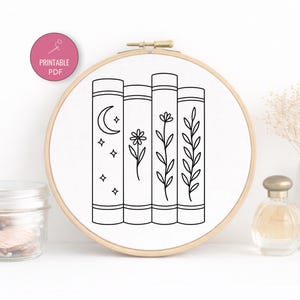 May include: A white fabric embroidery hoop featuring a line art design of four stacked books. The books are decorated with a moon and stars, a flower, and botanical elements. The text "PRINTABLE PDF" is in a pink circle.