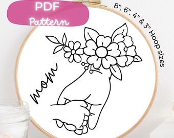 May include: Black and white embroidery pattern of a hand holding a smaller hand with the word "mom" and a floral design. The pattern is available in 8, 6, 4, and 3 inch hoop sizes.