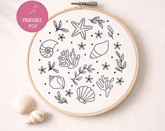 May include: A white embroidery hoop with a black line drawing of seashells, starfish, coral, and foliage. The text "PRINTABLE PDF" is in a pink circle. Two seashells are next to the hoop.