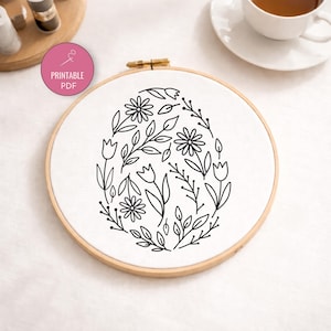May include: An embroidery hoop with a floral design in black line art, shaped like an egg. The design of flowers and leaves is on white fabric, held in a wooden hoop. A pink circle with "Printable PDF" text is visible.
