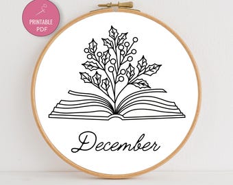 May include: A wooden embroidery hoop with a white fabric featuring a black line drawing of an open book with holly and berries, and the word "December". A pink circle with "Printable PDF" is in the upper left.