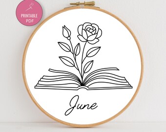 May include: A wooden embroidery hoop with a white fabric featuring a black line drawing of a rose growing from an open book. The word "June" is written below the book in cursive. A pink circle with "PRINTABLE PDF" is in the upper left.