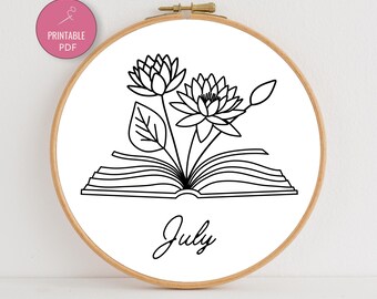 May include: A round embroidery hoop with a white background featuring a black line drawing of water lilies emerging from an open book. The word "July" is written below the book. A pink circle with "Printable PDF" is in the upper left.