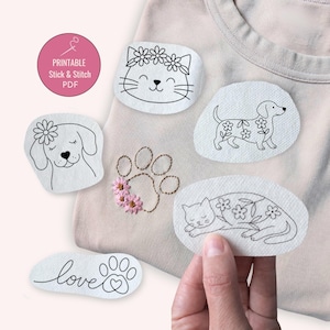 May include: A light beige t-shirt with several embroidered patches featuring line drawings of a cat, dog, and paw prints. The patches are white with black outlines and floral accents. The text "Printable Stick & Stitch PDF" is in a pink circle.