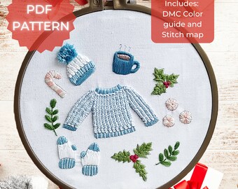 May include: Embroidered hoop art with a winter theme. The design includes a blue sweater, a white hat with a pom-pom, a candy cane, a cup of hot chocolate, holly leaves, and mittens.  Includes: DMC Color guide and Stitch map.