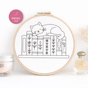 Puede incluir: Un bastidor de bordado en blanco y negro con un gato durmiendo sobre una pila de libros. Los libros están decorados con motivos florales y de hojas. Un círculo rosa con el texto "Printable PDF" está en la esquina superior izquierda.