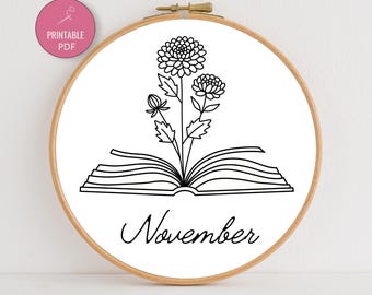 May include: A wooden embroidery hoop with a white fabric featuring a black line drawing of flowers emerging from an open book. The word "November" is written below the book in cursive. A pink circle with the words "PRINTABLE PDF" is in the upper left corner.