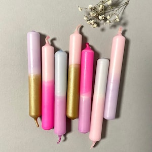 Könnte beinhalten: Eine Sammlung von acht farbigen Stabkerzen. Die Kerzen sind in Rosa-, Lila- und Weißtönen gehalten und haben goldfarbene Metallbasen. Einige Kerzen haben einen Farbverlauf. Ein Zweig getrockneter Blumen befindet sich im Hintergrund.