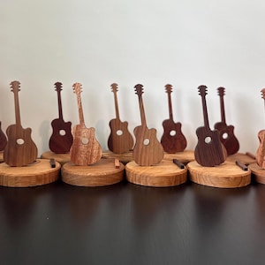 Puede incluir: Una fila de diez soportes de guitarra de madera, cada uno con un patrón de veta de madera diferente. Los soportes están dispuestos en una línea sobre una superficie oscura.