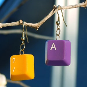 May include: A pair of earrings made from repurposed computer keys. One earring is yellow with a question mark and forward slash, the other is purple with the letter A.