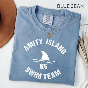 May include: A blue jean coloured t-shirt featuring the text "Amity Island Swim Team 1975" in white. The design includes a shark fin and waves. A gold necklace with a pendant is draped across the shirt.