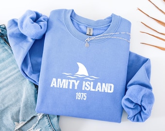 May include: A light blue sweatshirt with the text "AMITY ISLAND 1975" and a shark fin graphic. A silver necklace with a pendant is draped over the sweatshirt. Faded blue jeans are visible on the left side of the image.