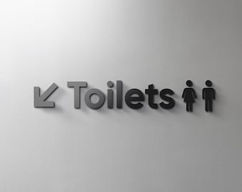 Puede incluir: Un letrero en blanco y negro con una flecha apuntando hacia abajo y la palabra "Toilets" en mayúsculas. También hay dos figuras de palo, una femenina y una masculina, a la derecha de la palabra "Toilets".