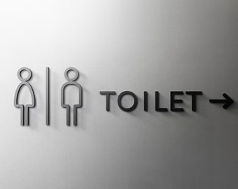 Può includere: Un cartello grigio per i servizi igienici con simboli femminili e maschili, una linea verticale, la parola "TOILET" e una freccia che punta a destra. Il cartello è montato su un muro grigio chiaro.