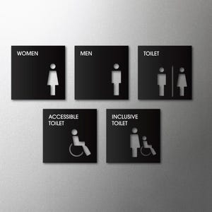 May include: Five black square restroom signs with white cut-out icons. The signs are labelled "WOMEN," "MEN," "TOILET," "ACCESSIBLE TOILET," and "INCLUSIVE TOILET." The signs are mounted on a grey wall.