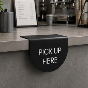 Puede incluir: Un letrero de mostrador de metal negro mate con un borde inferior curvo que cuelga sobre el borde de una superficie. El letrero presenta el texto blanco PICK UP HERE impreso en una fuente sans-serif clara, indicando una zona.