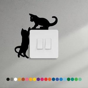 May include: A white light switch with two black cat silhouettes on it. The cat on top is sitting on the switch, while the cat on the bottom is reaching up towards the top cat.