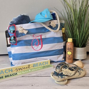 May include: A beach-themed still life featuring a blue and white striped tote bag with a pink initial, sunglasses, a blue cap, and sunscreen. Decorative wooden signs read "Beach Sweet Beach" and "At The Beach". Flip-flops and a potted plant complete the scene.