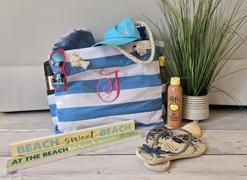 May include: A beach-themed still life featuring a blue and white striped tote bag with a pink initial, sunglasses, a blue cap, and sunscreen. Decorative wooden signs read "Beach Sweet Beach" and "At The Beach". Flip-flops and a potted plant complete the scene.