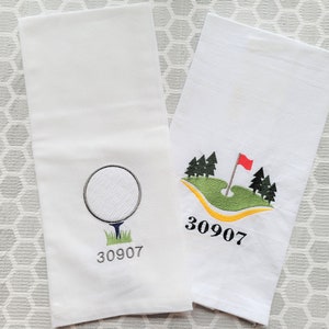 May include: Two white kitchen towels with embroidered golf-themed designs. One towel features a golf ball on a tee with the number 30907. The other towel features a green golf course with a flag and trees, and the number 30907.