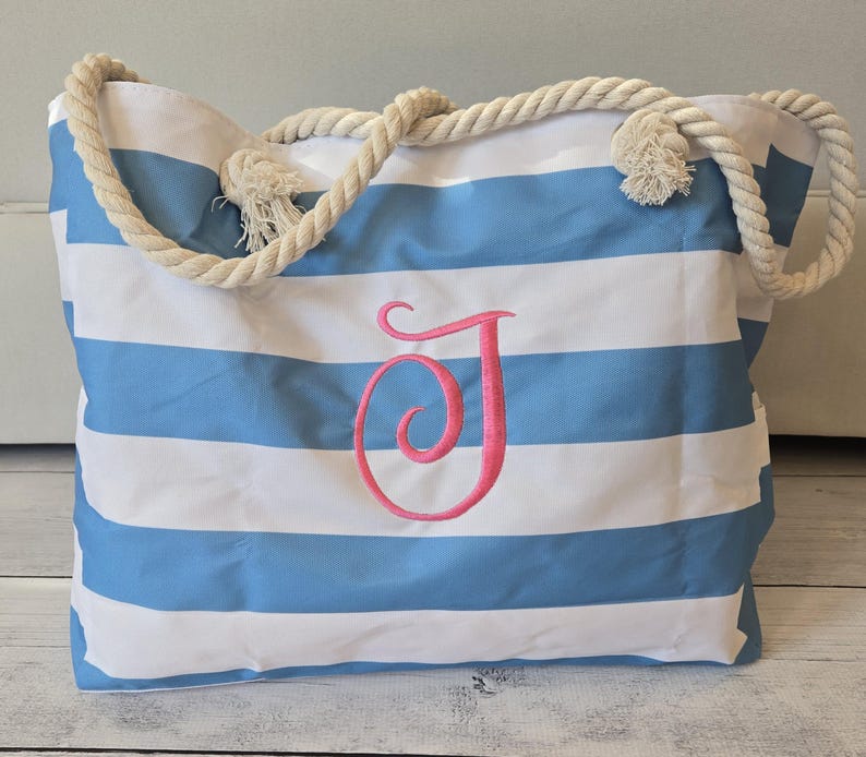 May include: A large beach tote bag with blue and white horizontal stripes. The bag features a pink embroidered letter "J" and sturdy rope handles. The bag is designed for carrying beach essentials.